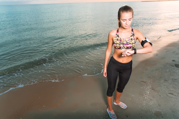 Muchacha apta de los jóvenes que mira el reloj durante ejercicios de la mañana en salida del sol de la playa