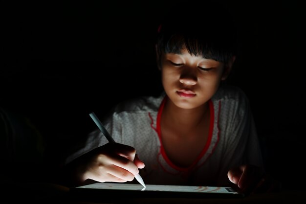 Muchacha asiática adolescente que usa el dibujo de la tableta mientras que miente en cama