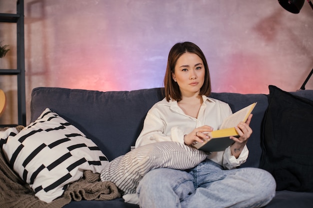 Foto muchacha asiática joven que lee un libro. la niña está sentada en el sofá. habitación cómoda.