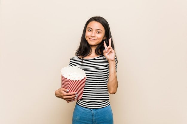 Muchacha asiática joven que sostiene un cubo de las palomitas que muestra el número dos con los dedos.