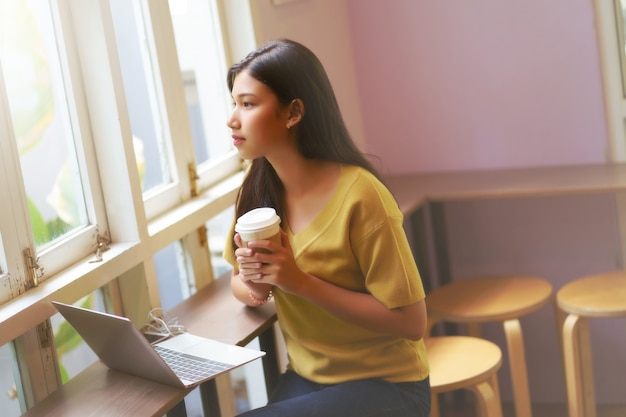 La muchacha asiática joven que sostiene la taza de papel y bebe el café caliente, utiliza la computadora portátil y la relajación