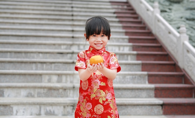 La muchacha asiática linda del niño en chino tradicional se viste con sostener la naranja sagrada en el templo chino