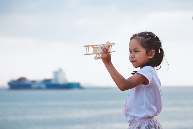 Muchacha asiática linda del niño que juega con el aeroplano de madera del juguete en la playa con felicidad