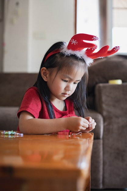 Muchacha asiática linda del niño que lleva el traje de la Navidad que ensarta perlas en una cuerda con intención y diversión en casa.