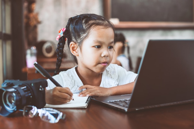 Muchacha asiática linda del niño que usa el ordenador portátil y escribiendo en su cuaderno en el café