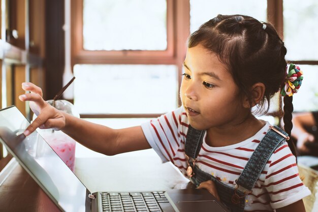 Muchacha asiática linda del niño que usa y que juega en el ordenador portátil en el café con la diversión y la felicidad