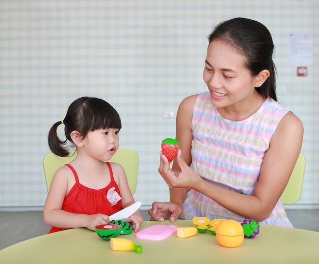 Muchacha asiática del niño y madre que juegan frutas plásticas en la habitación del niño