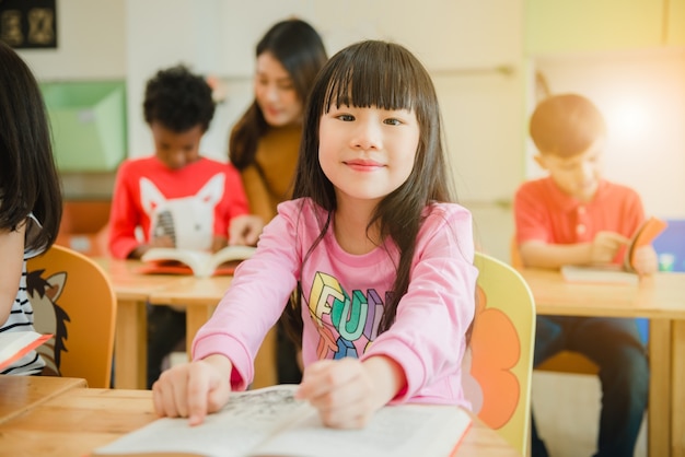 Muchacha asiática que lee un libro que sonríe en la cámara. Fila del libro de lectura elemental multiétnico de los estudiantes en sala de clase en la escuela. Imágenes de estilo de efecto vintage.