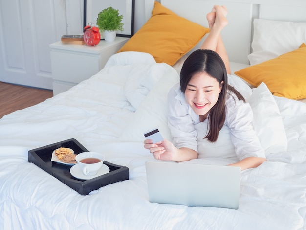 Muchacha asiática que usa la computadora con la tarjeta de crédito en cama