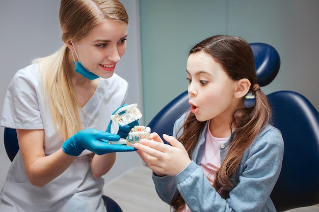 La muchacha asombrada emocionada se sienta en silla dental en el cuarto y toca la dentadura con los dientes artificiales. Dentista femenina sostenga y sonríe a la niña.