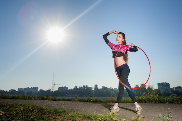 Muchacha atlética hermosa joven que se coloca con el aro del hula al aire libre en parque de la ciudad el día soleado.