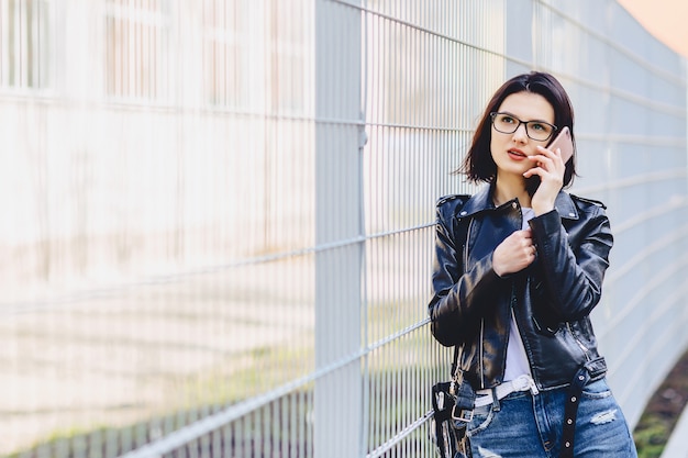 Muchacha atractiva en la chaqueta de cuero en vidrios que habla en el teléfono