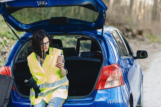 Muchacha atractiva que habla por teléfono cerca del coche en el camino en la chaqueta de seguridad