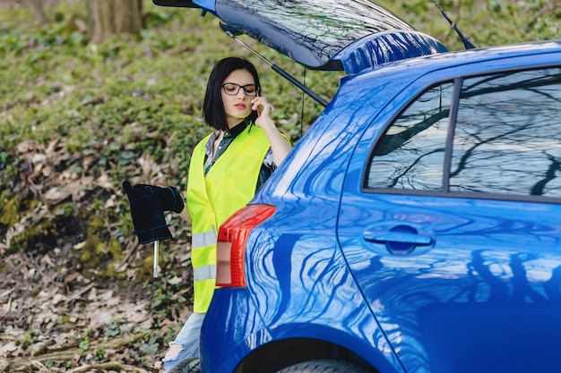 Muchacha atractiva que habla por teléfono cerca del coche en el camino en la chaqueta de seguridad