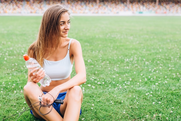 Muchacha bastante deportiva que se relaja en la hierba después de entrenar al aire libre con la botella de agua