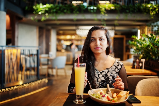 La muchacha bastante india en el vestido negro de la sari posó en el restaurante, sentado en la mesa con jugo y ensalada.