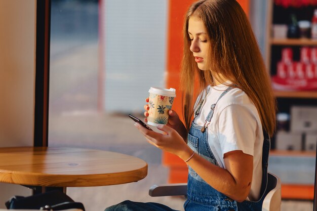 La muchacha bonita atractiva joven en el café con café y el teléfono en la mañana emite