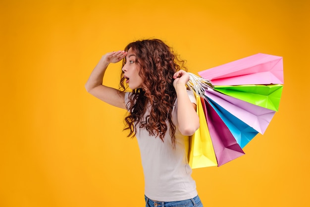 Muchacha bonita con los bolsos de compras que buscan y que buscan para las tiendas aisladas sobre amarillo