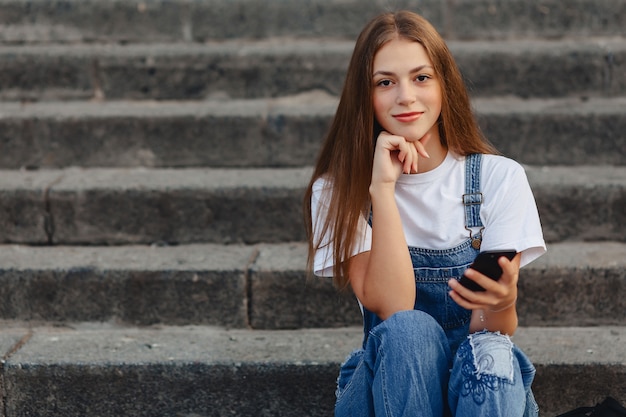 Muchacha bonita joven con la cartera que se sienta en las escaleras y que escribe SMS en el teléfono