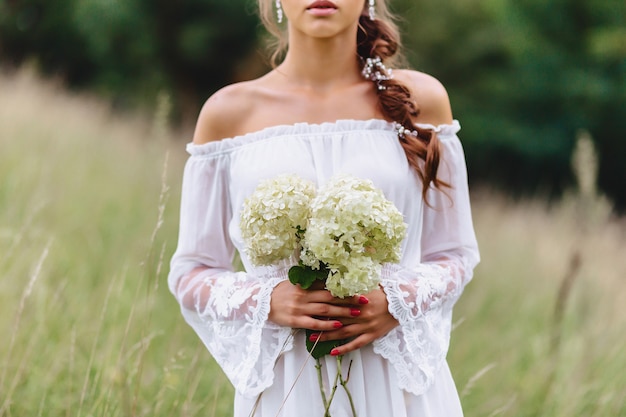 Muchacha bonita joven con la flor en paseo ligero del vestuario en el césped cerca del bosque del verano