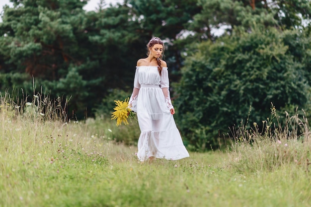 Muchacha bonita joven con la flor en paseo ligero del vestuario en el césped cerca del bosque del verano