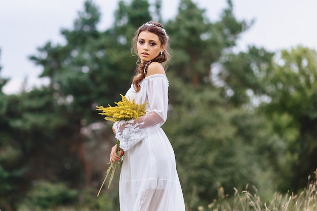 Muchacha bonita joven con la flor en paseo ligero del vestuario en el césped cerca del bosque del verano