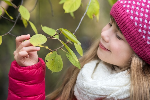 Muchacha bonita del niño que lleva la ropa caliente del invierno que sostiene la rama de árbol con las hojas verdes en tiempo frío al aire libre.