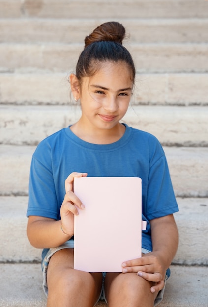 Foto muchacha del cabrito en la camiseta beige que se sienta en un banco y que sostiene el cuaderno en las manos de cerca, tiro recortado. maqueta de cuaderno