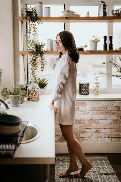 La muchacha en una camisa blanca bebe el café por la mañana en una cocina.