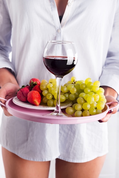 Foto la muchacha en una camisa sostiene una bandeja de fruta y de vino en el fondo blanco