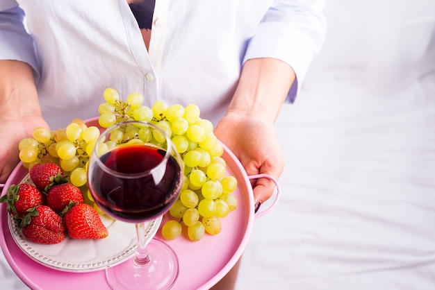 La muchacha en una camisa sostiene una bandeja de fruta y de vino en el fondo blanco