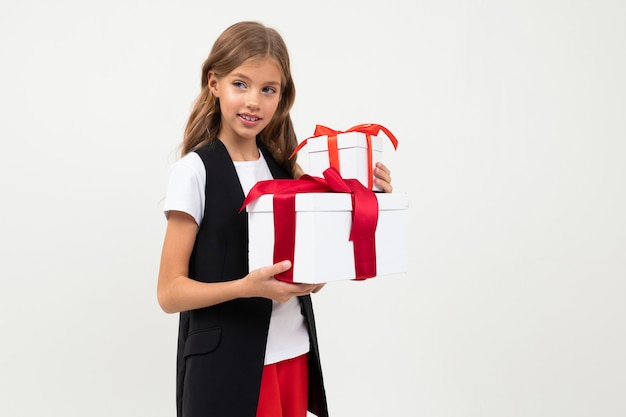 La muchacha caucásica del adolescente en la chaqueta negra, la camisa blanca y los pantalones rojos sostiene cajas blancas con los regalos y se regocija aislado en blanco