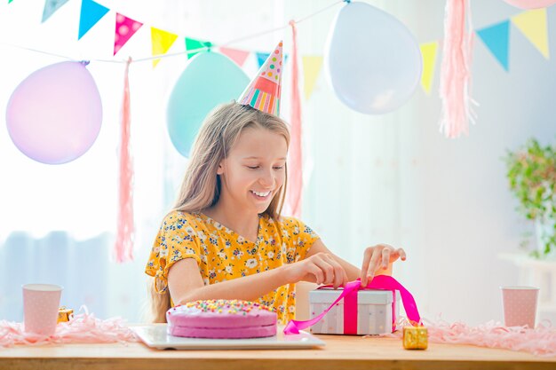 La muchacha caucásica está soñadoramente sonriendo y mirando el pastel de cumpleaños del arco iris. Fondo colorido festivo con globos. Concepto de fiesta y deseos de cumpleaños.