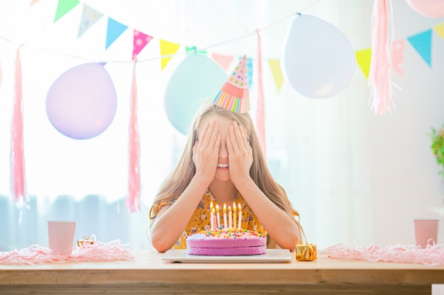 La muchacha caucásica está soñadoramente sonriendo y mirando el pastel de cumpleaños del arco iris. Fondo colorido festivo con globos. Concepto de fiesta y deseos de cumpleaños.