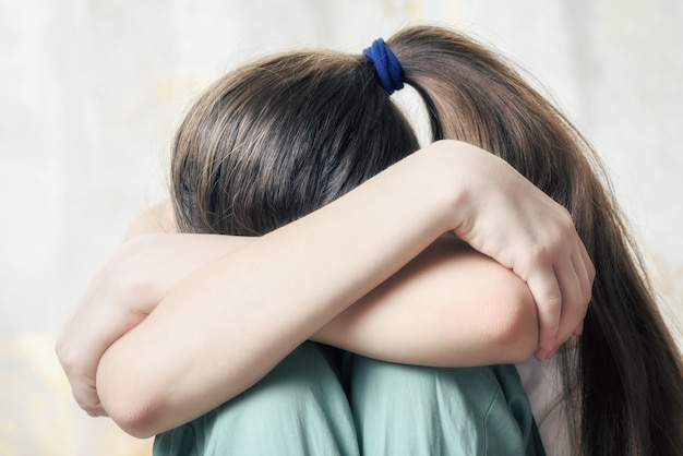 La muchacha con la coleta larga se sienta acurrucada apretando sus rodillas con sus manos. El concepto de abuso infantil.