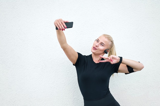 Muchacha deportiva atractiva joven que lleva la ropa de deportes negra que escucha la música y que estira en el fondo blanco de la pared. Concepto de vida activa y saludable.