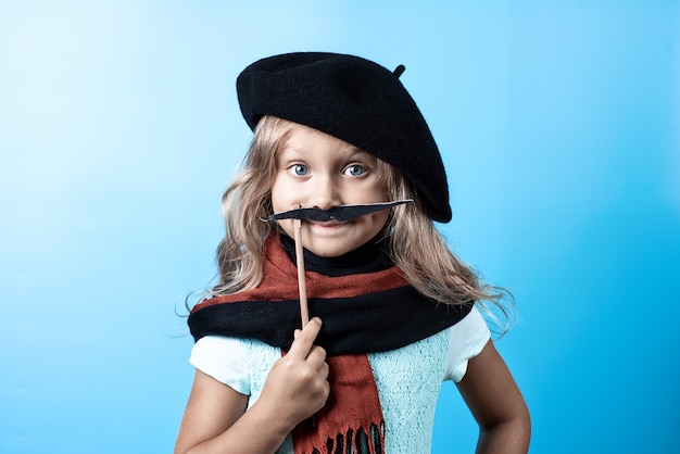 Muchacha divertida en boina negra, bufanda y bigote en un palo en azul