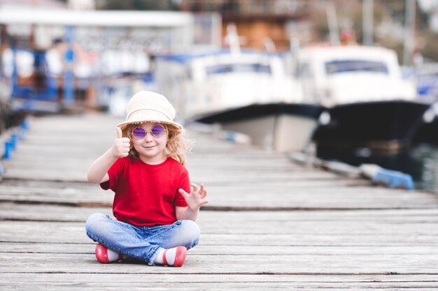 Muchacha divertida del niño que se sienta en el muelle del mar use ropa casual