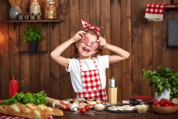 Foto muchacha divertida que cocina la pizza y que engaña con las rebanadas de salami