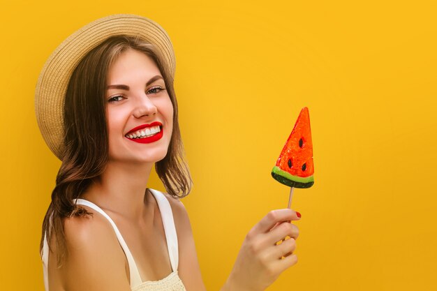 Muchacha elegante joven en un sombrero de paja con una piruleta multicolora en un fondo amarillo