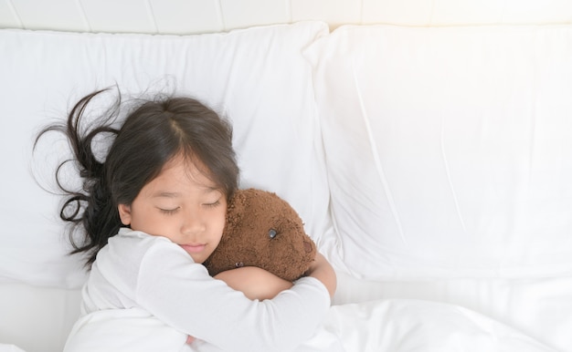 La muchacha está abrazando el oso de peluche mientras que duerme en cama
