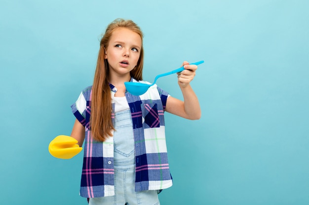 La muchacha europea sonriente sabe con una cuchara en sus manos en la pared azul clara