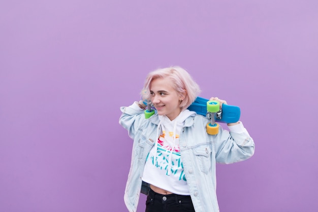 La muchacha feliz se coloca con una patineta contra el fondo de una pared púrpura y sonríe. Elegante chica rubia con una patineta sobre un fondo morado.