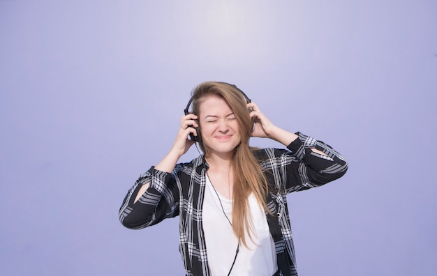 La muchacha feliz emocional con los auriculares en su cabeza se coloca en un fondo púrpura, brillante y escucha la música. Estudiante en ropa casual escucha música en el fondo de una pared de color púrpura.