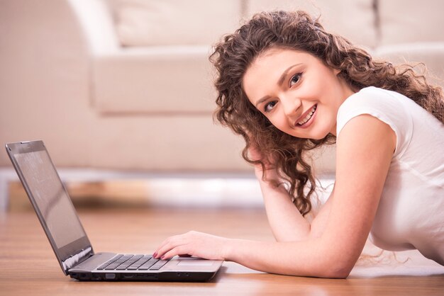 La muchacha feliz está tendida en el suelo y está trabajando en una computadora portátil.