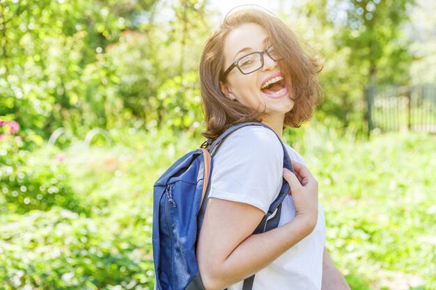 Muchacha feliz hermosa del estudiante positivo en lentes con la mochila que sonríe en parque verde. Mujer descansando en el campus durante el almuerzo. Concepto de educación y ocio.