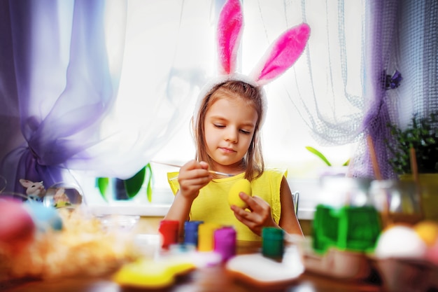 Muchacha feliz de pascua en orejas de conejo que pinta los huevos, niño pequeño en casa. vacaciones de primavera