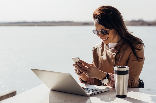 Foto muchacha feliz que manda un sms en un teléfono elegante afuera en día asoleado brillante.