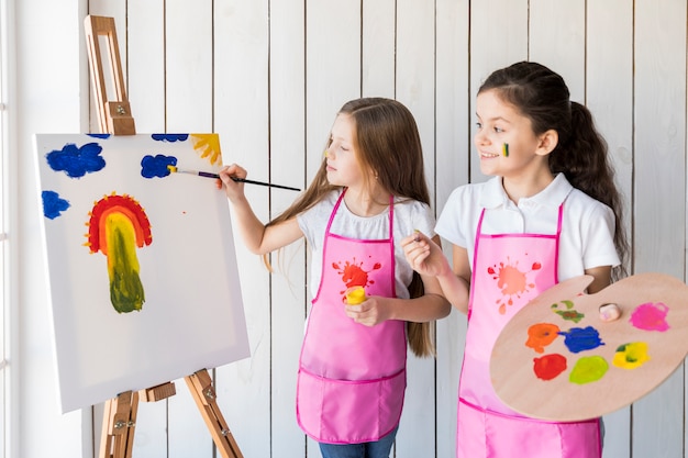 Foto muchacha feliz que sostiene la paleta en la mano que mira a su amigo que pinta en la base con el cepillo de pintura