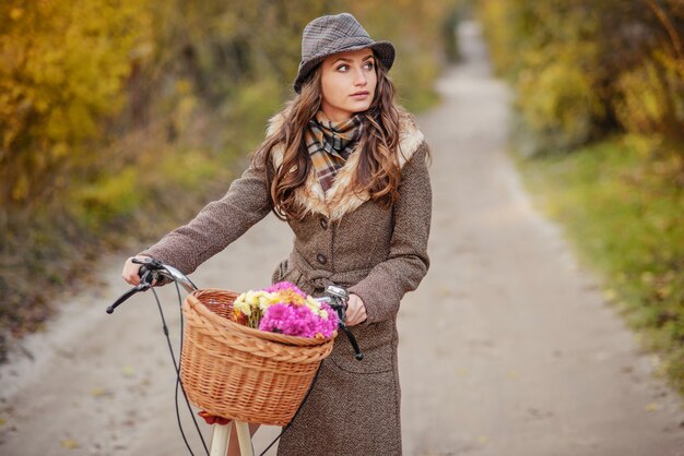 La muchacha hermosa en abrigo y el sombrero marrones del otoño que llevan a las señoras monta en bicicleta con la cesta llena de flores y de árboles del otoño en el fondo.